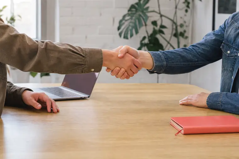 Twee mensen geven elkaar een hand boven een lichte vergadertafel met een laptop